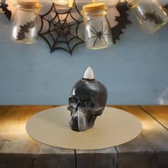 Dark glass skull with a white cone top, smoke emanating from its eye sockets and nose, placed on a round beige surface; spooky Halloween-themed decoration at Monalisa Medical Store.