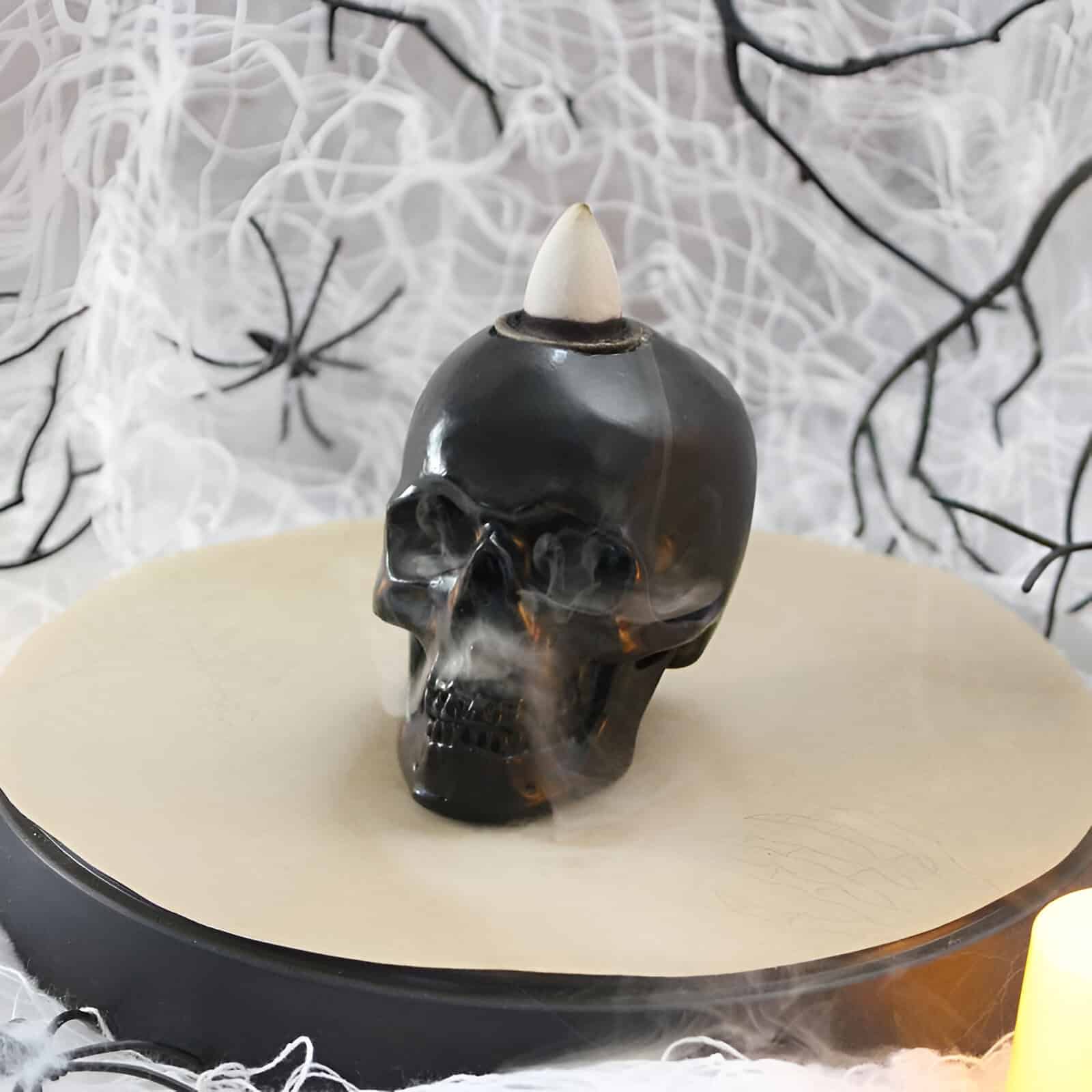 Black skull-shaped Halloween decoration with fog smoke effect, topped with a white horn, on a reflective surface with spider web background.