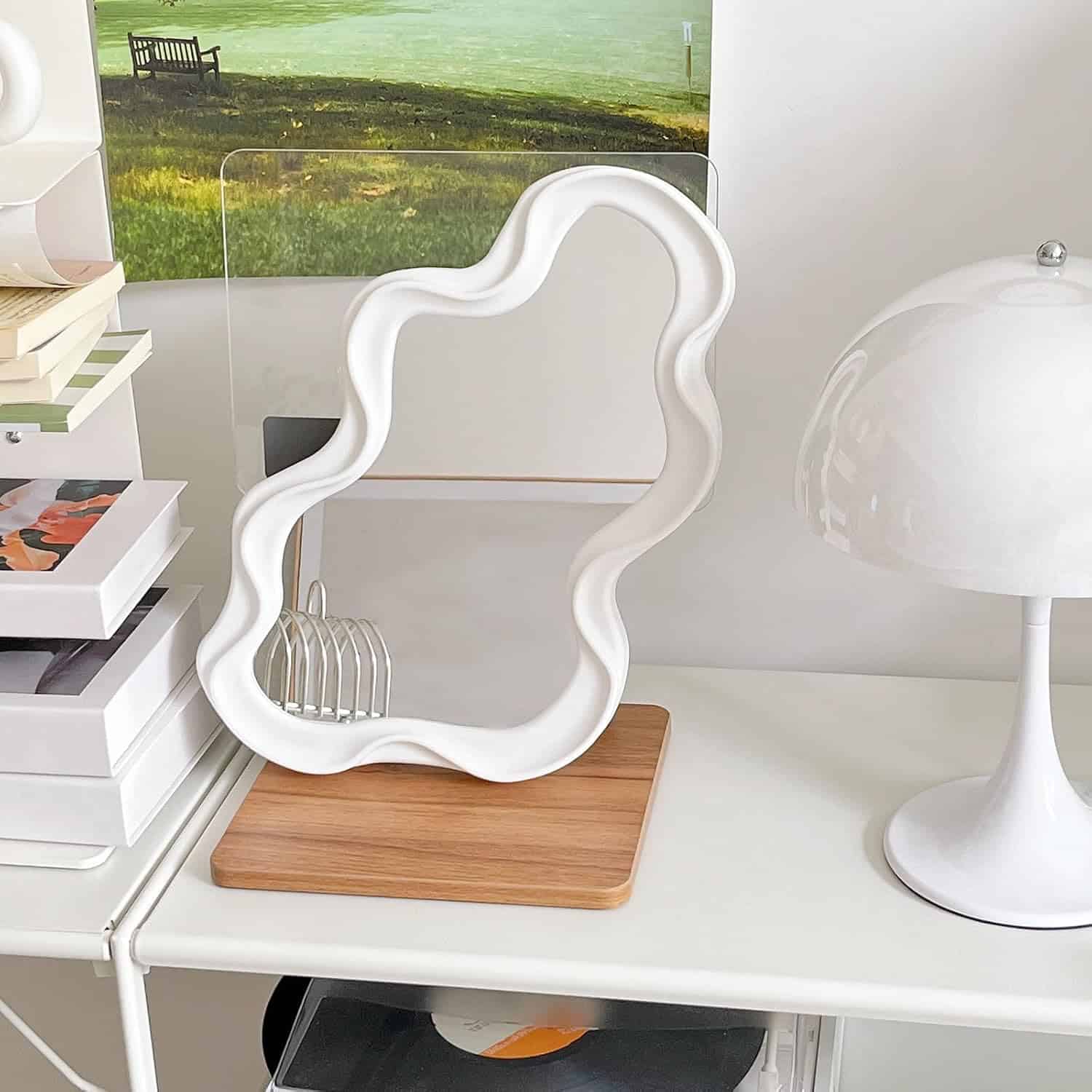Elegant white wavy-edge mirror with wooden base, modern white table lamp, and stacked fashion books in a stylish workspace, emphasizing contemporary decor and aesthetic appeal for interior design or lifestyle SEO.