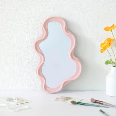 Reflective pink wavy-shaped vanity mirror on white wall, beauty and skincare accessories on table.