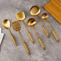 Shiny gold kitchen utensils set including slotted spoon, serving spatula, ladle, pasta server, and tongs on grey surface. Perfect for modern cooking and stylish kitchen design.