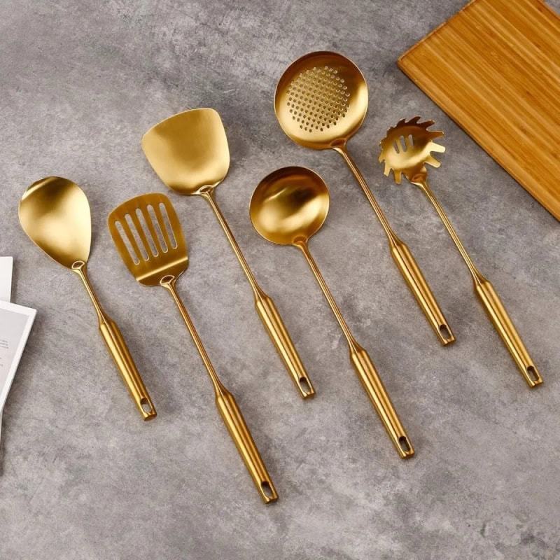 Shiny gold kitchen utensils set including slotted spoon, serving spatula, ladle, pasta server, and tongs on grey surface. Perfect for modern cooking and stylish kitchen design.