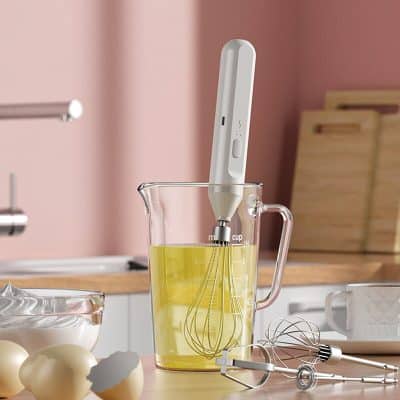 Whisk and electric hand mixer in a measuring cup with eggs and baking utensils on a pink kitchen countertop.