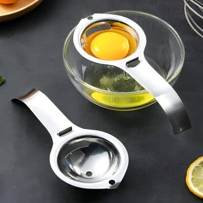 Stainless steel egg separator placed over a glass bowl with a raw egg, kitchen utensils and lemon slices on a black surface.