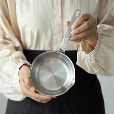 Stainless steel saucepan held by a person in a cream blouse, ideal for cooking or kitchen use.