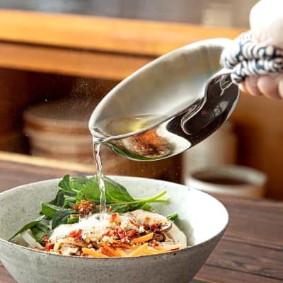 Fresh greens and pasta topped with cheese and vegetables in a bowl, with a hand pouring olive oil from a ladle, emphasizing healthy eating and gourmet food presentation.