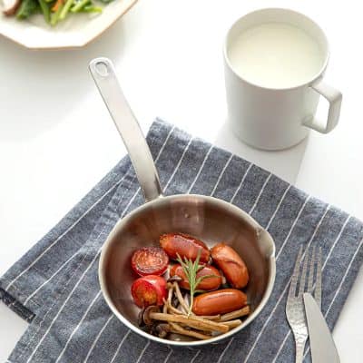 Fresh cooked sausages, cherry tomatoes, and mushrooms in a stainless steel pan served with milk in a white mug, on a striped cloth napkin, representing healthy meal options at Monalisa Store.