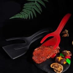 Black and red silicone tongs with grilled steak and colorful appetizers on a black slate serving platter, set against a dark background with green fern leaves for stylish food presentation.