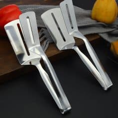 Stainless steel kitchen spatulas on a dark surface with apples and a wooden cutting board.