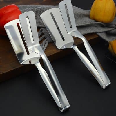 Stainless steel kitchen spatulas on a dark surface with apples and a wooden cutting board.