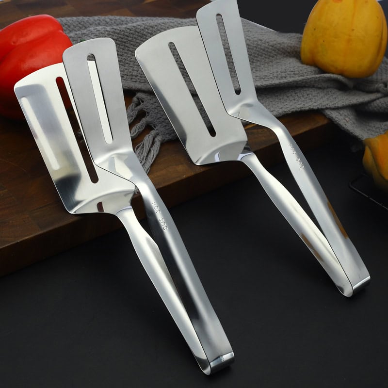 Stainless steel kitchen spatulas on a dark surface with apples and a wooden cutting board.