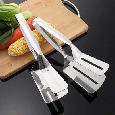 Stainless steel kitchen tongs on black surface with fresh vegetables including corn, carrot, and bok choy.