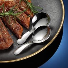 Premium stainless steel teaspoon set on a black skillet with cooked meat, highlighting elegant cutlery for dining and kitchen essentials.
