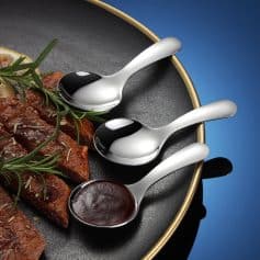 Stainless steel soup spoons on a black plate with grilled meat and herbs, ideal for serving and enjoying delicious meals at Monalisa Store.