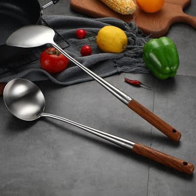 Stainless steel kitchen ladles with wooden handles on gray surface with fresh vegetables and kitchen accessories.