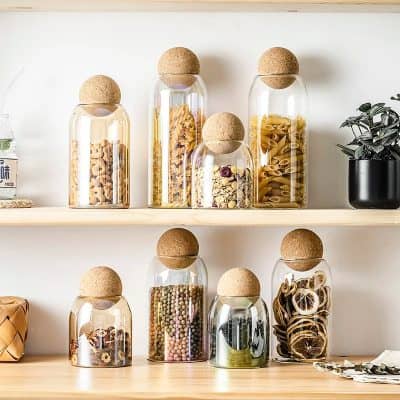 Cereal storage jars with cork lids on wooden shelves, showcasing various pasta shapes, dried herbs, and snacks in a modern, organized kitchen display.