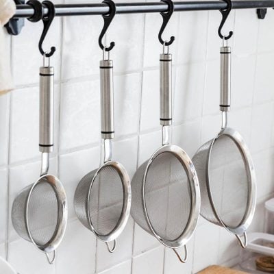 Stainless steel mesh strainers hanging on a kitchen rack against white tiled wall.
