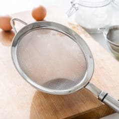 Fine mesh kitchen sieve or strainer on wooden cutting board for cooking or baking, with eggs and baking supplies in background.