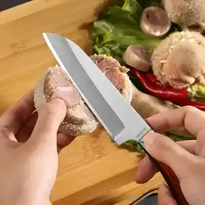 Fresh frozen chicken thigh being cut with a sharp kitchen knife on wooden cutting board with vegetables in the background.