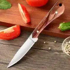 Sleek kitchen knife with wooden handle on rustic wooden surface, surrounded by fresh tomatoes, green chili, and white peppercorns, ideal for cooking and food preparation.