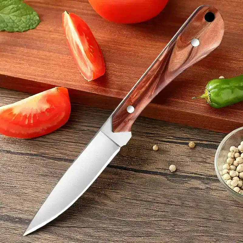 Sleek kitchen knife with wooden handle on rustic wooden surface, surrounded by fresh tomatoes, green chili, and white peppercorns, ideal for cooking and food preparation.