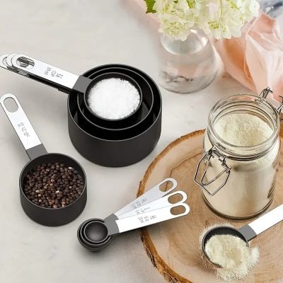 Soft-close measuring spoons, salt and pepper grinders, and a jar of protein powder arranged on a wooden tray with a flower vase and pink cloth. Ideal for kitchen measurements and cooking prep.