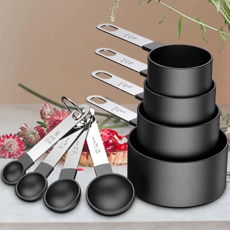 Black measuring cups and spoons set for kitchen and culinary measurements, stacked and laid out with measurement labels, ideal for cooking and baking, displayed on a table with a cake and flowers in the background.