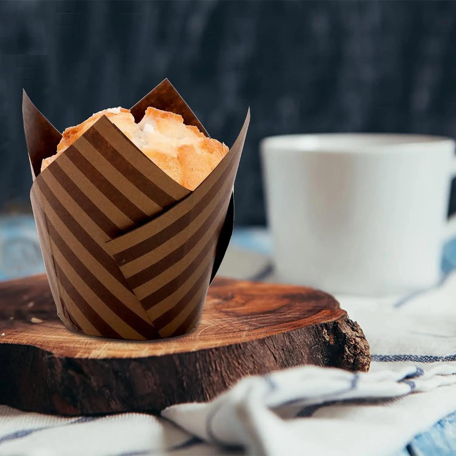 Freshly baked muffin wrapped in striped paper, placed on a wooden serving board with a white coffee cup in the background. Perfect for a cozy morning or a coffee break.