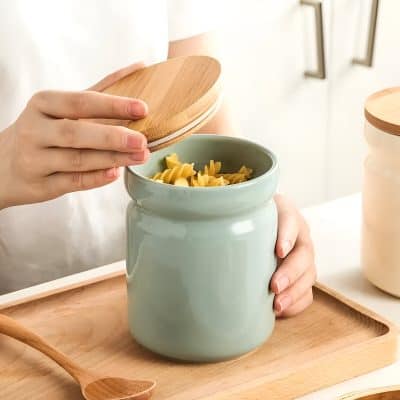 Fresh fusilli pasta being stored in a light green ceramic jar with a wooden lid, on a wooden tray, emphasizing kitchen storage solutions and pasta organization for home cooking.