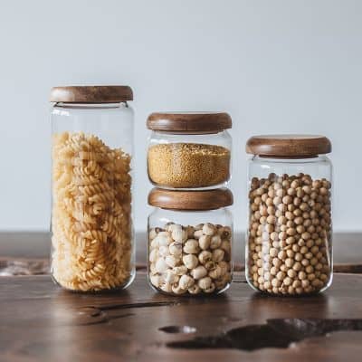 Glass jars with wooden lids filled with pasta, grains, and legumes on a rustic wooden surface, showcasing organized storage solutions for a healthy kitchen.
