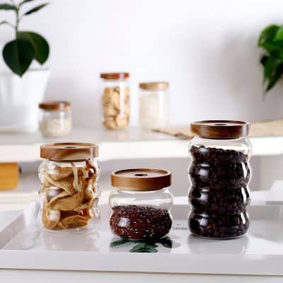 Glass jars with wooden lids containing coffee beans and dried herbs on a white tray in a modern, minimalist kitchen setting.