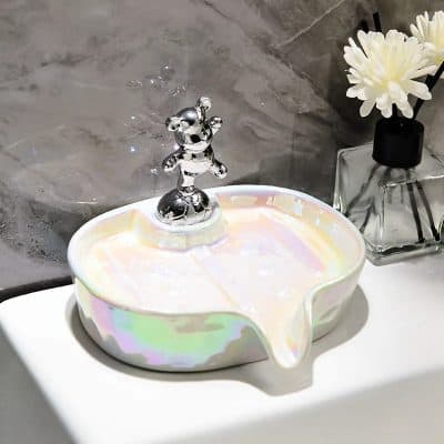 Shiny silver faucet on iridescent soap dish with flow design, modern bathroom decor, elegant tile background, and a glass vase with white flowers in the background.