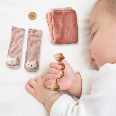 Cute baby socks with cat face design, soft peach-colored blanket, baby wooden rattle, and a small round wooden toy on white background at Monalisa Store.