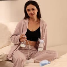 Alt text: Woman using breast pump for lactation at home, relaxed in casual pink pajamas, sitting on bed, focusing on breastfeeding support with modern medical equipment.