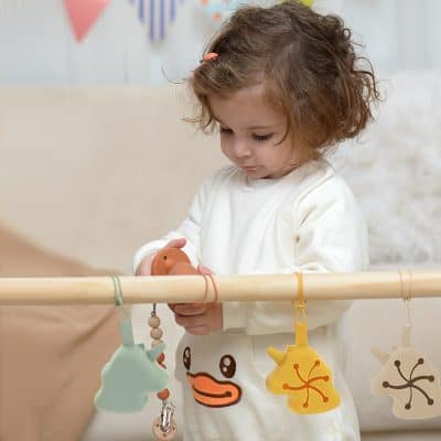 Soft baby girl playing with teething toys hanging on a wooden rail at Monalisa Medical Store, focusing on infant health and baby teething accessories.