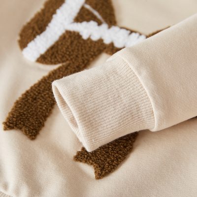 Soft beige sweatshirt with ribbed cuff and brown and white textured patch logo for casual fashion. Perfect for comfortable, stylish everyday wear and urban street style.