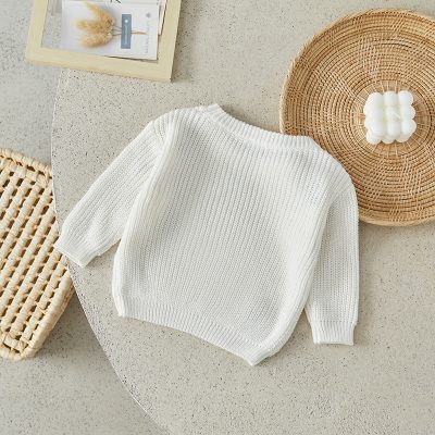 Soft white knitted sweater displayed on a neutral textured surface, accented with wicker baskets, a framed botanical print, and calming candles, perfect for stylish and cozy wardrobe essentials.