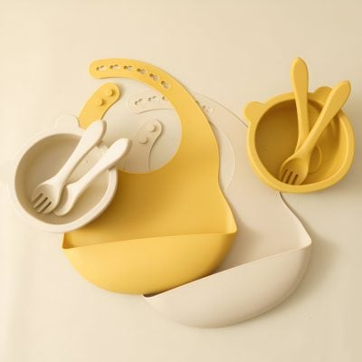 Colorful silicone baby dish set with fork and spoon in yellow and cream. Perfect for independent eating, featuring a bib-shaped tray with nested bowls and utensils designed for children.