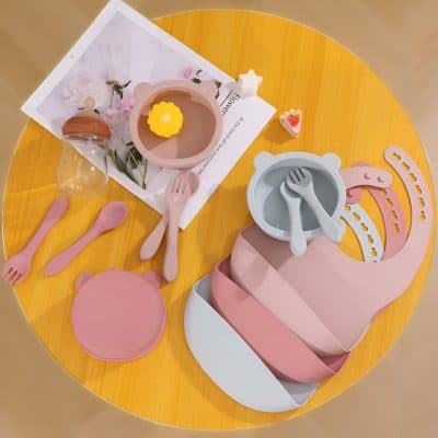 Pink and gray silicone children's dishware set with utensils, bowls, and plates on a yellow table, perfect for kids' mealtime and fun dining experiences.
