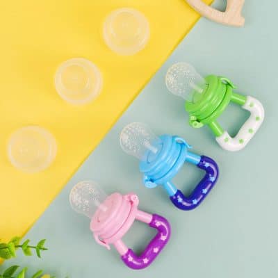 Pink, blue, and green baby pacifiers with silicone nipples on pastel yellow and blue background, emphasizing baby care and oral health supplies.