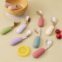 Colorful ergonomic baby spoons with soft silicone handles and stainless steel scoops, perfect for feeding infants, displayed on a pastel background with natural wooden bowl, lemon slice, and cherries.