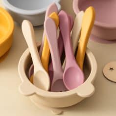 Pink, beige, and yellow silicone spoons in a white bowl, perfect for baby feeding; colorful and safe for infants.