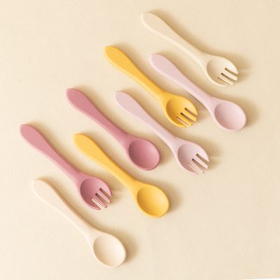 Colorful silicone baby spoons in pastel pink, yellow, beige, and lavender for feeding and toddler dining, arranged on a soft beige background.