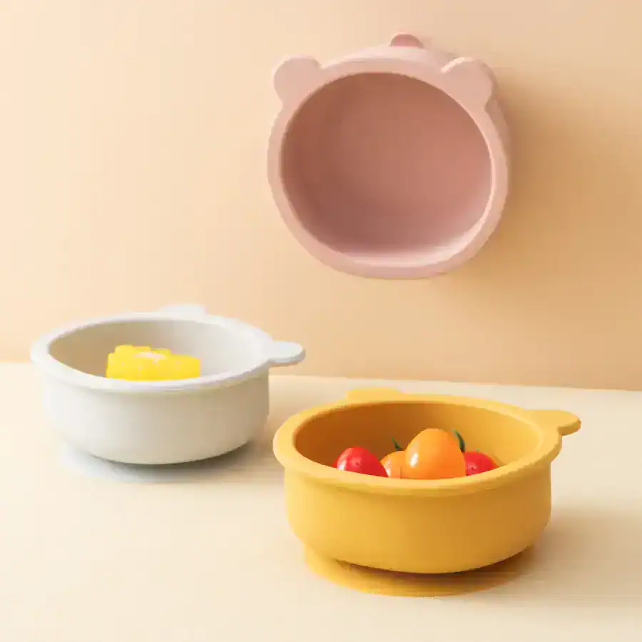 Brightly colored bear-shaped silicone bowls, one yellow filled with small tomatoes and a white one with butter, placed on a clean white surface with a pastel pink bear-shaped lid hanging on the wall.