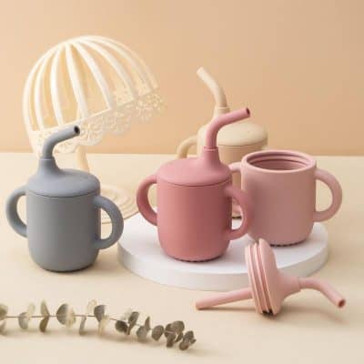 Gray baby sippy cup with soft silicone straw and handles for toddlers, pastel pink and cream sippy cups, and a silicone lid, on a white round platform with a decorative lamp in the background, ideal for kids' drinkware and preschooler accessories.