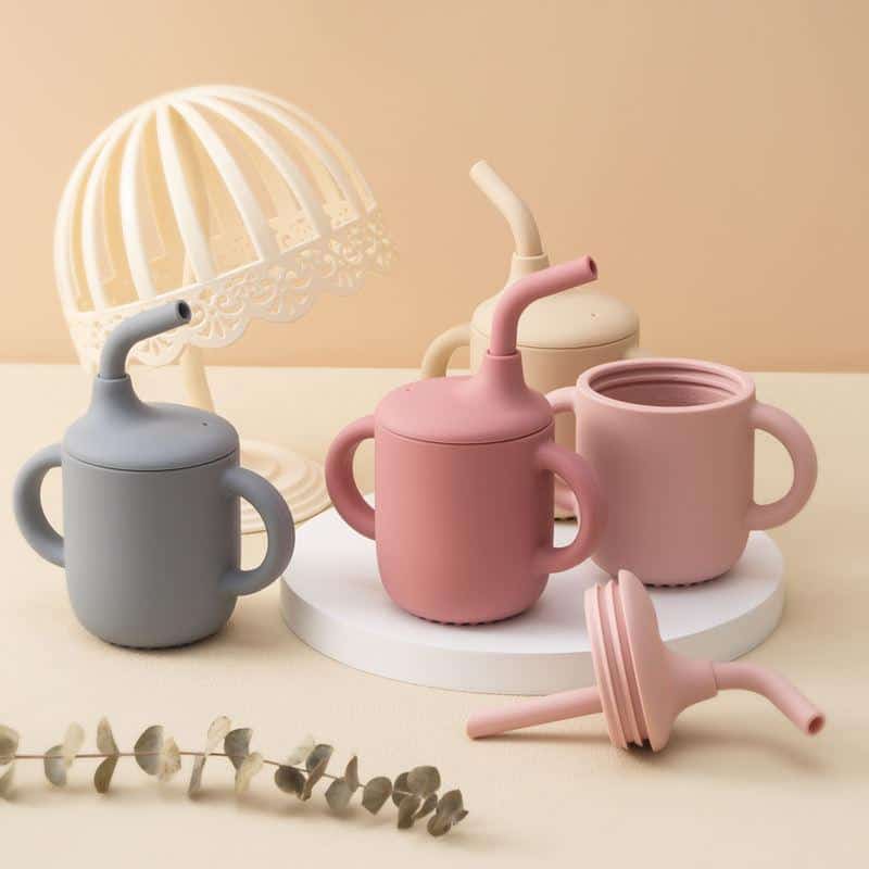 Gray baby sippy cup with soft silicone straw and handles for toddlers, pastel pink and cream sippy cups, and a silicone lid, on a white round platform with a decorative lamp in the background, ideal for kids' drinkware and preschooler accessories.