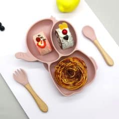 Colorful kid's silicone plate with divided sections for desserts and pasta, featuring pink animal paw design, with child-friendly utensils and a yellow apple in the background.