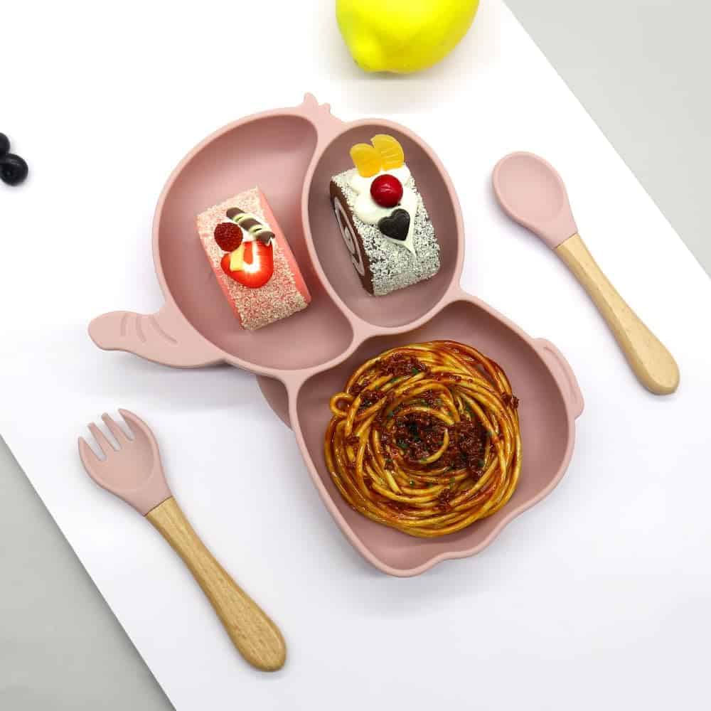 Colorful kid's silicone plate with divided sections for desserts and pasta, featuring pink animal paw design, with child-friendly utensils and a yellow apple in the background.