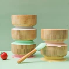 Wooden ice cream bowls stacked with pastel-colored silicone spoons, on a green background with a tomato, showcasing eco-friendly food serving options from Monalisa Store.