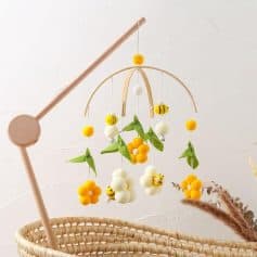Beaded baby crib mobile with yellow, white, and green felt flowers and bumblebee accents hanging above a woven bassinet, ideal for nursery decor and sensory stimulation.
